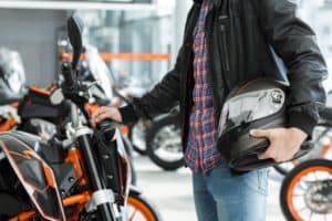 Man wearing leather jacket standing near his newly bought motorbike holding a black helmet.